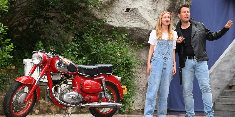 Zwei Theaterschauspieler stehen neben Motorrad auf einer Bühne