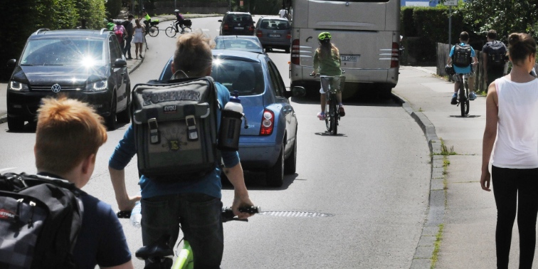 Schülerinnen und Schüler laufen auf Gehweg und fahren Fahrrad auf Straße