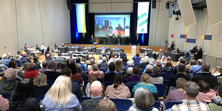 Versammlung in einem Saal mit Bühne, Podium und Videokonferenz auf großem Bildschirm.