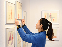 Kunst Eine Frau in blauem Pullover hängt in einer Galerie Bilder an die Wand. Die Bilder zeigen abstrakte Zeichnungen in hellen Farben.