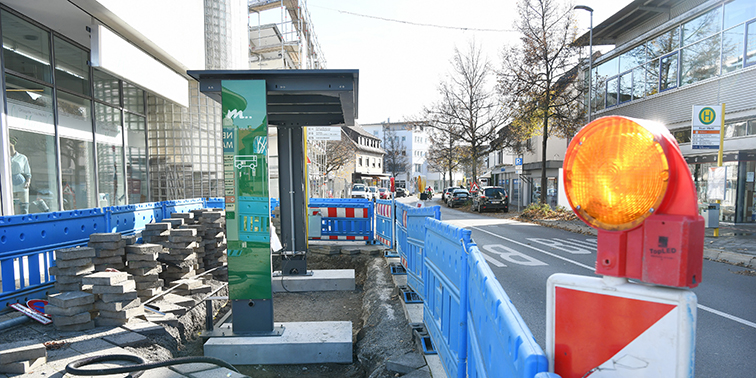 Baustelle einer Bushaltestelle mit Absperrungen und Warnleuchte, im Hintergrund eine Straße mit Gebäuden.