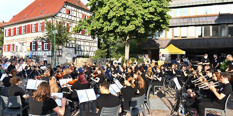 Musik hören, Musik machen! Ein großes Orchester spielt im Freien auf einem Platz, umgeben von Publikum. Die Musiker tragen schwarze Kleidung und spielen verschiedene Instrumente, darunter Geigen, Trompeten und Flöten. Im Hintergrund sind Fachwerkhäuser und weitere Gebäude zu sehen.